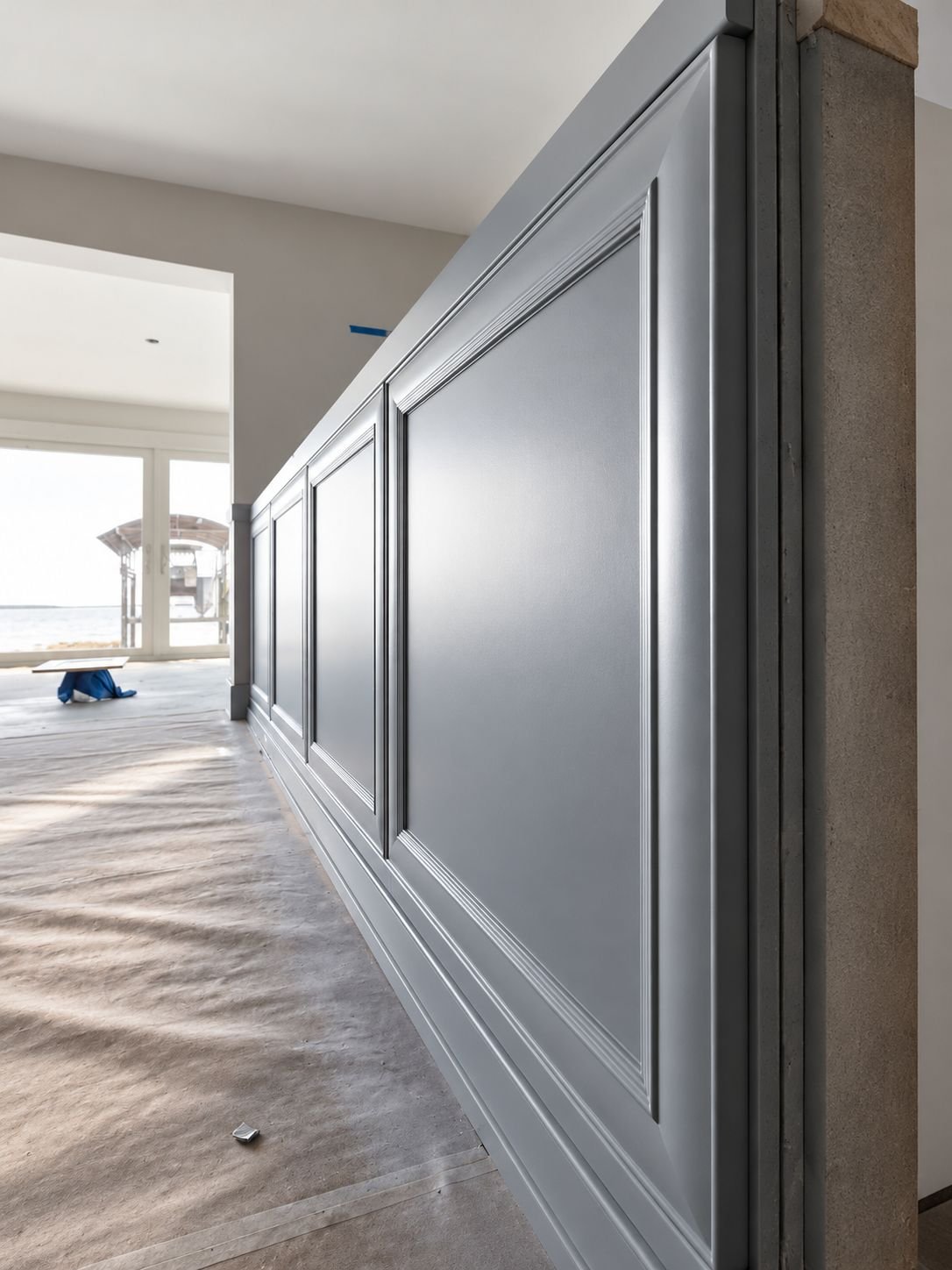 Long wall of gray raised-panel wainscoting in a bright home with protective paper on the floor